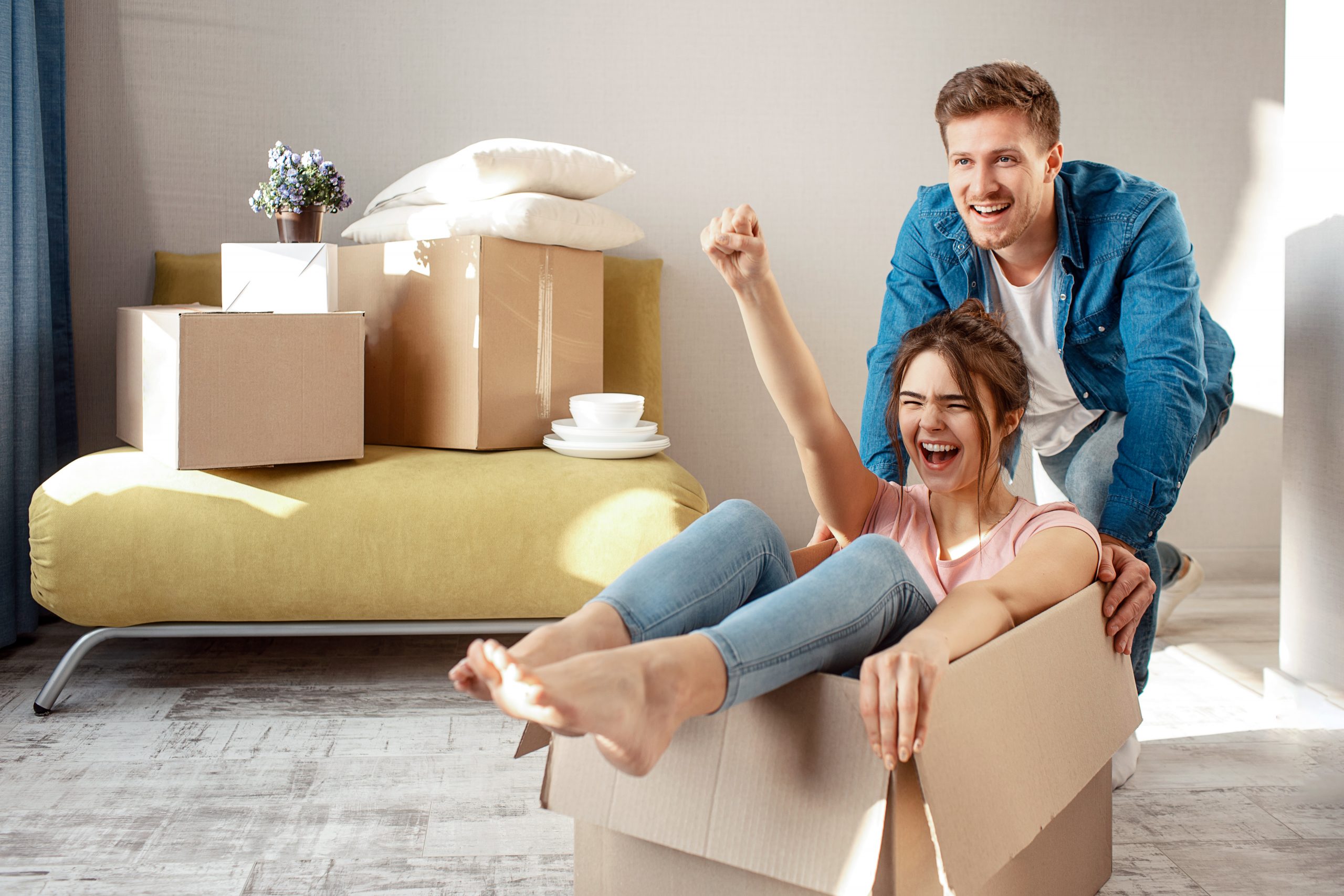 Young family couple bought or rented their first small apartment. Cheerful woman scream sitting in box. Guy move her. They play game during moving in